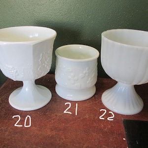 May include: Three white glass pedestal bowls. The bowl on the left has a hexagonal shape with a grape vine design. The middle bowl is cylindrical with a floral pattern. The bowl on the right has a fluted design.