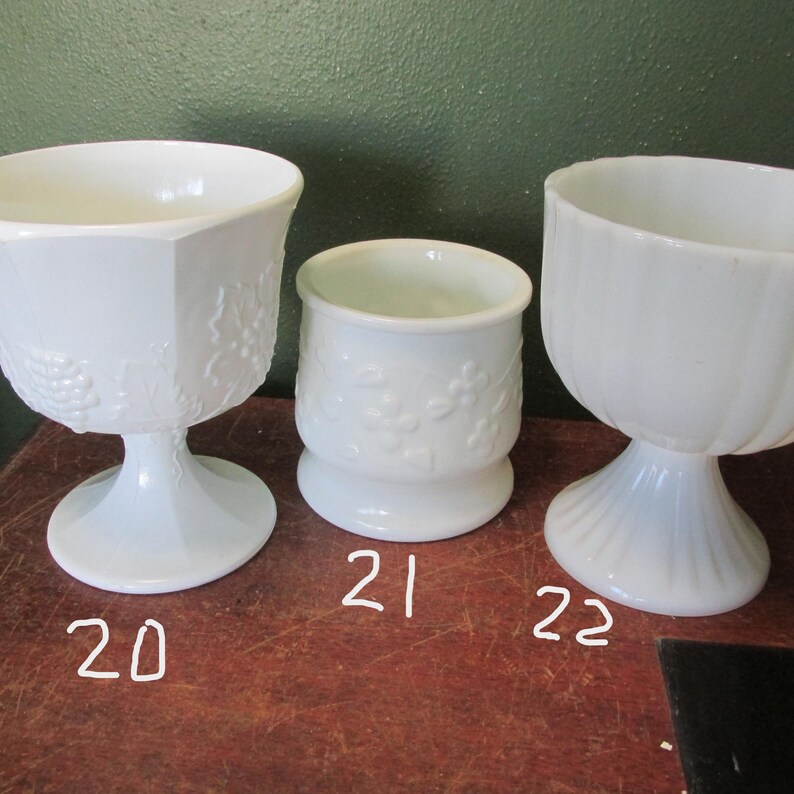 May include: Three white glass pedestal bowls. The bowl on the left has a hexagonal shape with a grape vine design. The middle bowl is cylindrical with a floral pattern. The bowl on the right has a fluted design.