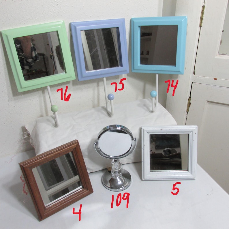 May include: A selection of decorative mirrors in different styles and colours. Features square mirrors with pastel green, blue, and white frames, a brown framed mirror, and a round, silver vanity mirror.