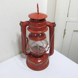 May include: A red metal kerosene lantern with a glass globe and a wire cage. The lantern has a handle on top and a carrying handle on the side.