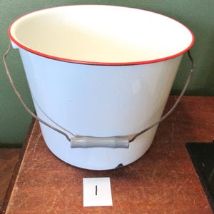 May include: White enamel bucket with a red rim and a wire handle with a wooden grip. The bucket is sitting on a wooden surface.