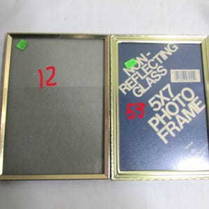 May include: Two gold-colored rectangular photo frames. One frame is empty, while the other contains a blue backing with the text "NON-REFLECTING GLASS 5X7 PHOTO FRAME". The frames are side-by-side, with the number "12" written in red on the empty frame.