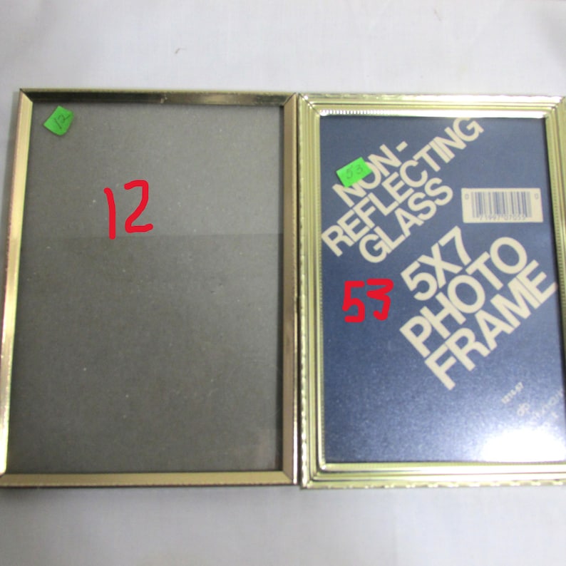 May include: Two gold-colored rectangular photo frames. One frame is empty, while the other contains a blue backing with the text "NON-REFLECTING GLASS 5X7 PHOTO FRAME". The frames are side-by-side, with the number "12" written in red on the empty frame.