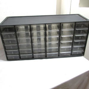 May include: A black storage cabinet featuring thirty individual clear plastic drawers arranged in a grid of six columns and five rows. Each drawer has a small integrated handle on the front, providing a view of the contents stored inside.