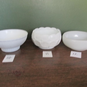 May include: Three white glass bowls of varying designs are displayed on a brown surface. The bowls include a scalloped edge bowl, a geometric patterned bowl, and a bowl with a textured exterior. Each bowl has a corresponding number on a small white tag.