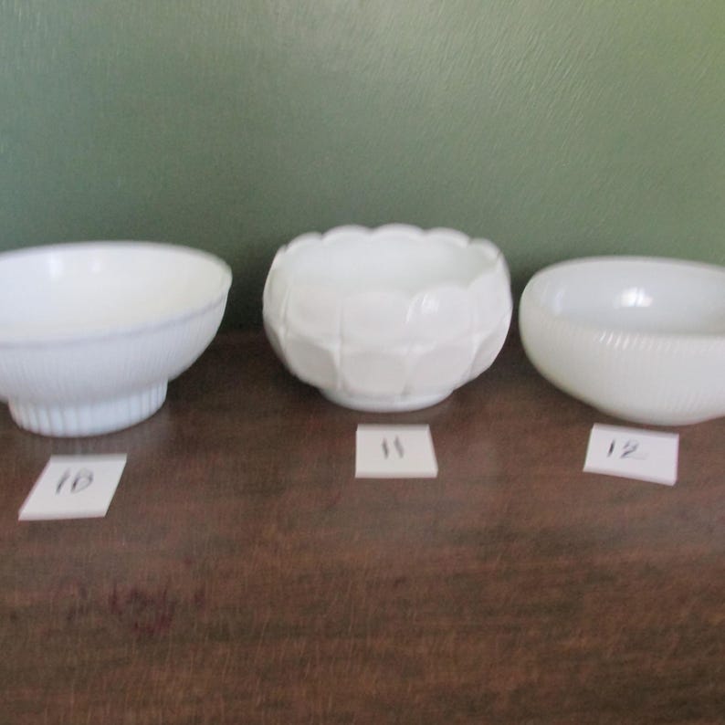 May include: Three white glass bowls of varying designs are displayed on a brown surface. The bowls include a scalloped edge bowl, a geometric patterned bowl, and a bowl with a textured exterior. Each bowl has a corresponding number on a small white tag.