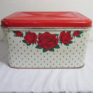 May include: A rectangular metal storage tin with a bright red lid and a cream-colored base. The body features a repeating pattern of small red and green diamond shapes and a central floral design of large red roses with green leaves.
