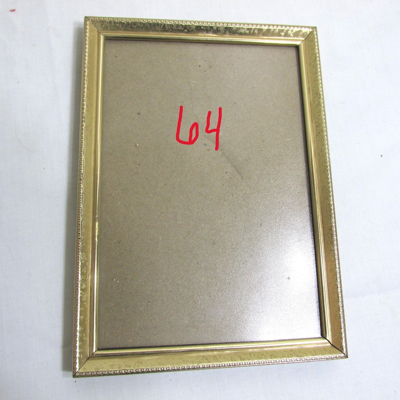 May include: A rectangular gold-colored picture frame with a textured border. The frame has a light brown backing and the number "64" is written in red in the center. The frame is empty.