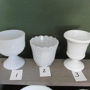 May include: Three white vintage glass containers. The containers include a pedestal bowl, a hobnail pot, and a small urn. Each container is on a wooden surface with a green background.