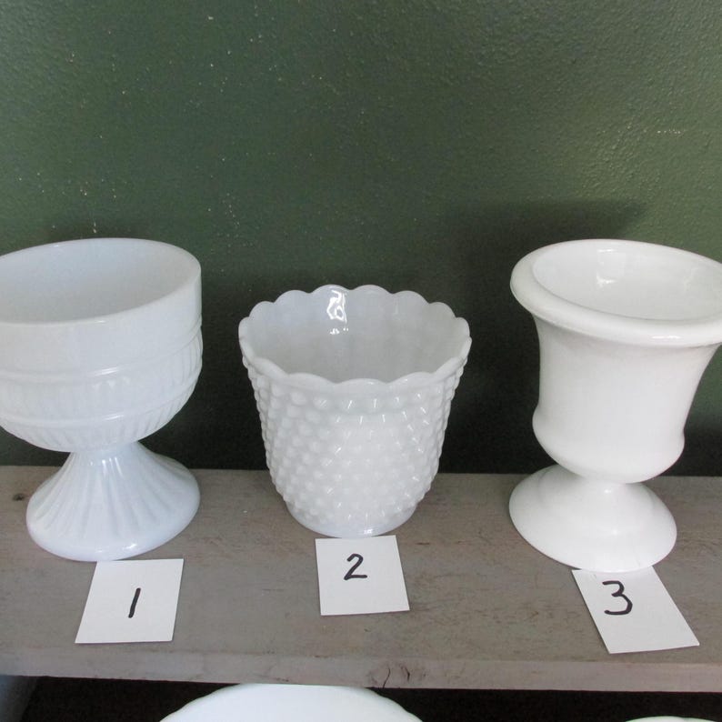 May include: Three white vintage glass containers. The containers include a pedestal bowl, a hobnail pot, and a small urn. Each container is on a wooden surface with a green background.
