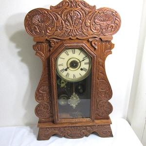 May include: A brown wooden mantel clock featuring intricate floral carvings on the frame. The clock face has a cream background with black Roman numerals and two hands. A glass panel reveals a gold-toned pendulum and internal clock mechanisms.