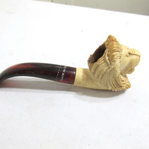 May include: A carved tobacco pipe featuring a lion's head design. The pipe has a curved stem with a dark red and brown finish, and a light-colored bowl. The text "MADE IN IT" is visible on the stem.