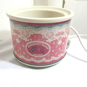 May include: A pink and white Rival Potpourri Crock slow cooker with a floral and lace design. The crock pot has a white interior and a white rim. The text "Potpourri Crock" and "RIVAL" are visible on the front.