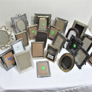 May include: A collection of 16 silver, gold, and black picture frames of various sizes and styles. The frames are arranged on a white surface.