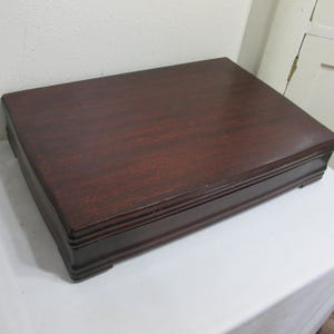 May include: A rectangular, dark brown wooden box with a slightly raised lid. The box has a smooth, polished surface with visible wood grain. The edges are subtly detailed, and the box sits on small feet. The overall design is simple and classic.