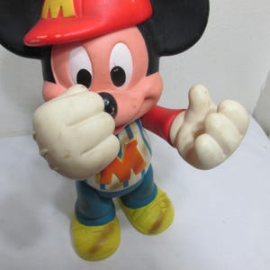 K&ouml;nnte beinhalten: Eine Vintage-Mickey-Mouse-Figur in Feuerwehruniform. Die Figur hat einen roten Hut mit einem gelben "M", ein blau-wei&szlig; gestreiftes Hemd mit einem gelben "M" und gelbe Schuhe. Die Figur befindet sich in einer spielerischen Pose.