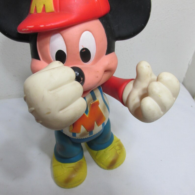 K&ouml;nnte beinhalten: Eine Vintage-Mickey-Mouse-Figur in Feuerwehruniform. Die Figur hat einen roten Hut mit einem gelben "M", ein blau-wei&szlig; gestreiftes Hemd mit einem gelben "M" und gelbe Schuhe. Die Figur befindet sich in einer spielerischen Pose.