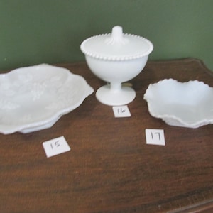 May include: Three white, vintage glass serving dishes on a wooden surface. The set includes a covered candy dish with a beaded rim, a scalloped-edge serving plate, and a small dish with a decorative edge. The background is a solid green.