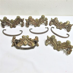 May include: A collection of antique brass drawer pulls and handles. The ornate pulls feature a gold finish with intricate floral and scroll designs. The handles are a curved, dark metal. The hardware is likely for a cabinet or dresser.