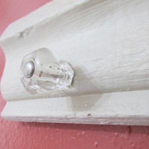 May include: A clear glass knob with a silver center is attached to a white painted wooden trim. The knob has a faceted design and is mounted on a light-colored, decorative molding against a pink background.