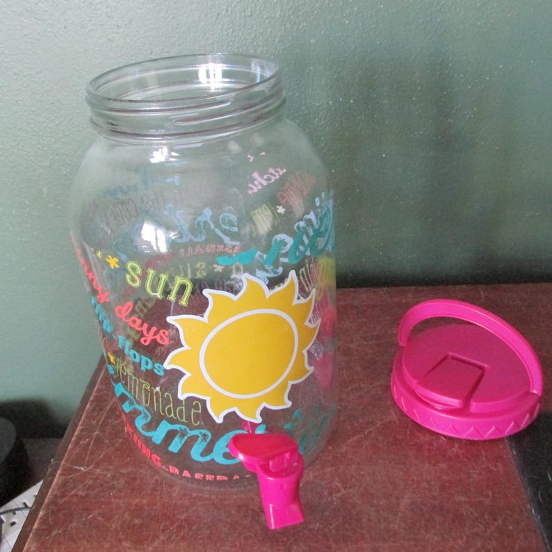 May include: A clear glass beverage dispenser featuring a large yellow sun graphic and colorful text including the words sun, days, and lemonade. The dispenser has a bright pink plastic spigot at the base and a matching detachable pink lid.