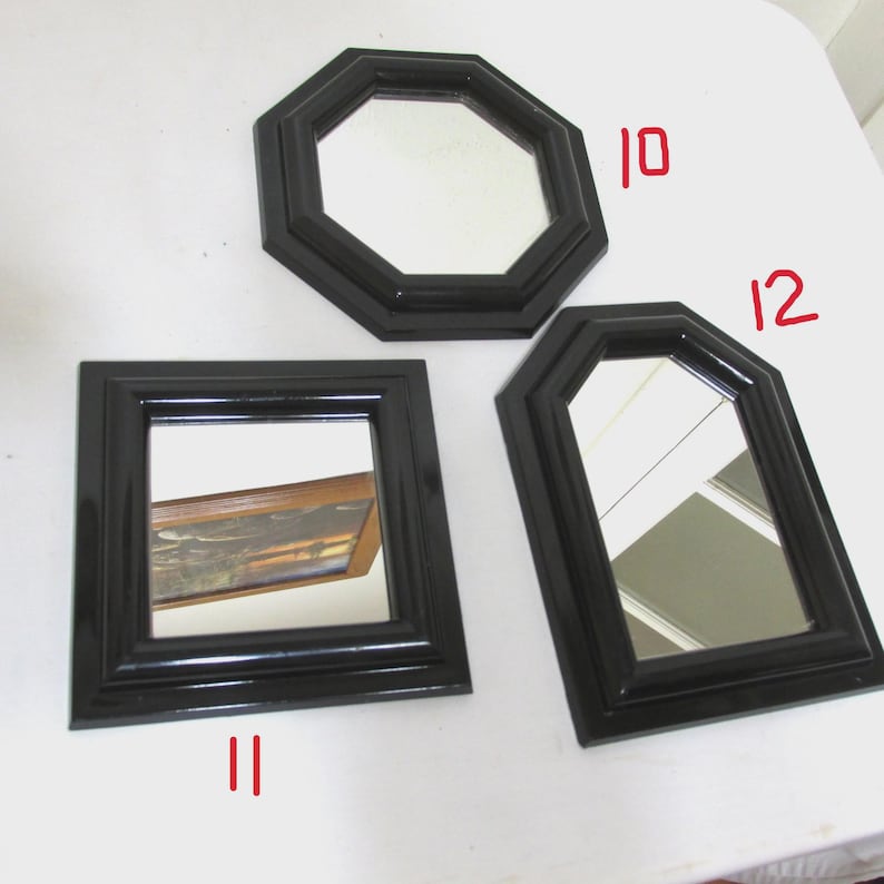 May include: Three small, black-framed mirrors in different geometric shapes: an octagon, a square, and a modified arch. The mirrors reflect the surrounding environment, and are displayed on a white surface.