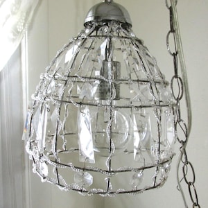 May include: A decorative pendant light with a silver metal frame and clear crystal accents. The light fixture has a rounded cage design, with a bulb visible inside. The crystals are various shapes and sizes, adding sparkle.