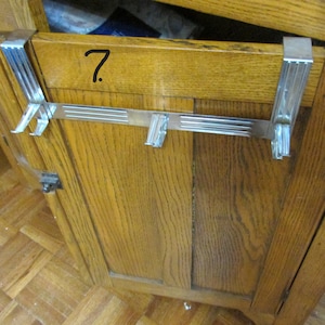 May include: A silver-colored metal over-the-door hanger with multiple hooks. The hanger is attached to a wooden cabinet door. The number 7 is written in black marker on the cabinet door.