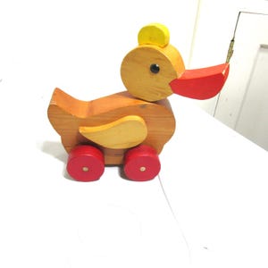 May include: A wooden pull-along toy duck with a yellow head crest and red beak and wheels. The duck's body is a light brown color, with lighter wood accents on the wings. The wheels are red. A classic children's toy.