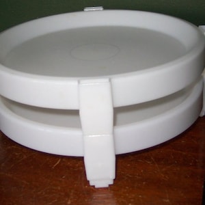 May include: A white plastic container with a lid and a handle. The container has a round shape with a flat top and a slightly raised rim. The handle is attached to the side of the container and is used to open and close it.