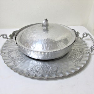 May include: A hammered aluminum serving dish with a lid and a decorative tray. The dish has a floral design on the lid and tray, with a scalloped edge. The lid has a tulip-shaped handle. The tray has handles on either side.