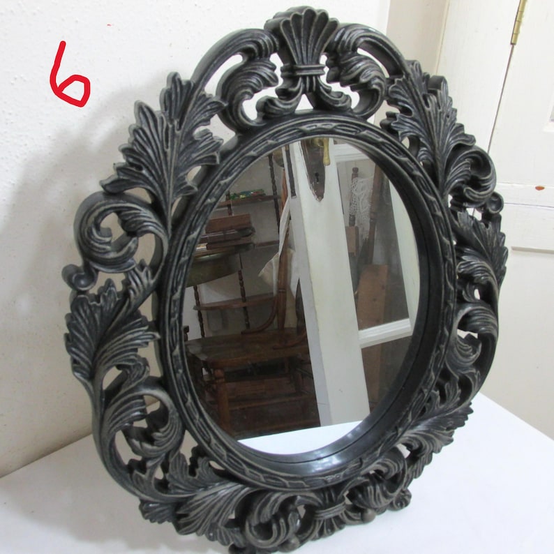 May include: Oval mirror with a dark grey, ornate frame. The frame has a detailed, carved leaf and scroll design. The mirror reflects the surrounding environment. A classic piece for home decor.