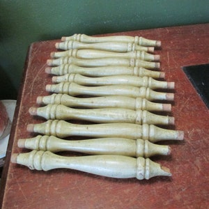 May include: A stack of 12 unfinished wooden chair legs, each with a rounded top and a slightly tapered bottom. The legs are a light beige color and have a smooth, sanded finish.