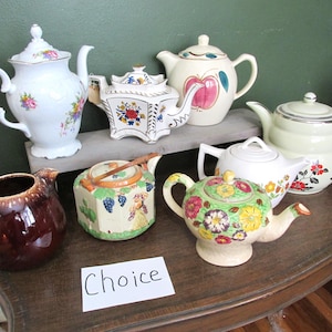 May include: A collection of vintage teapots in various shapes and sizes. The teapots feature floral, fruit, and geometric designs in a range of colors, including white, cream, brown, and green. A sign that says "Choice" is in the foreground.