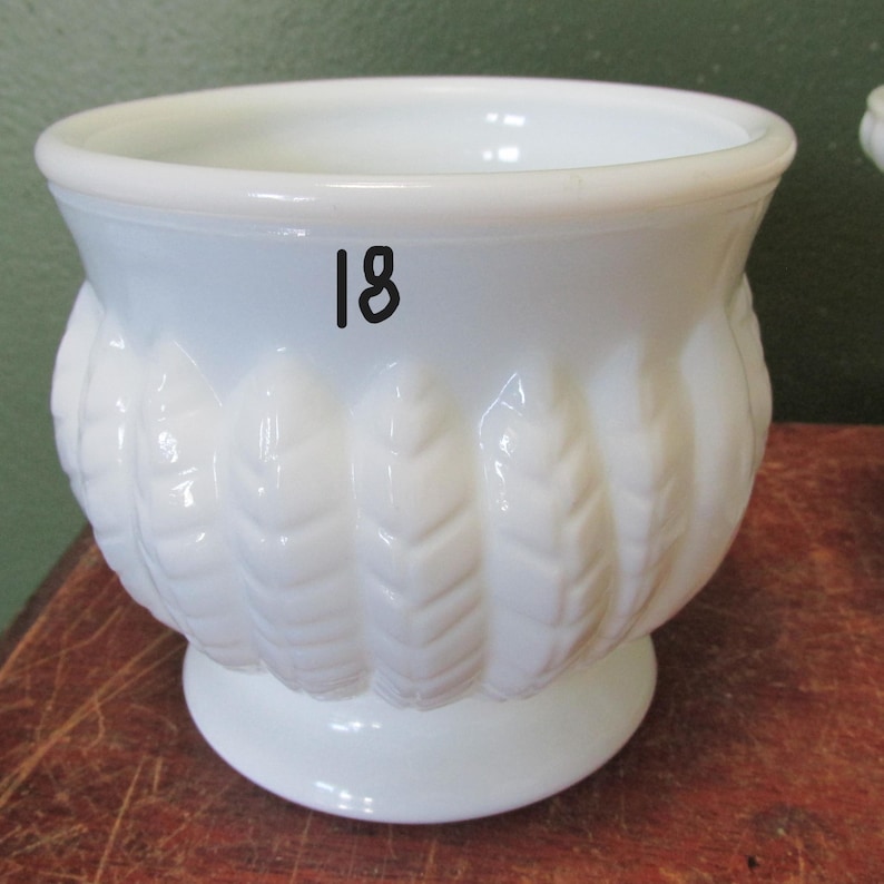 May include: A white, opaque glass planter with a pedestal base. The sides of the planter feature a repeating leaf design. The number "18" is printed in black on the side of the planter. The planter is sitting on a wooden surface.