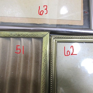 May include: A close-up of three framed pictures. The frames are gold and silver. The numbers 51, 62, and 63 are written in red on the pictures. The pictures are behind glass.