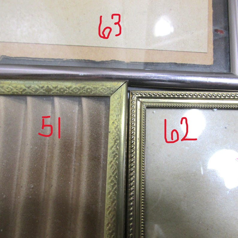 May include: A close-up of three framed pictures. The frames are gold and silver. The numbers 51, 62, and 63 are written in red on the pictures. The pictures are behind glass.