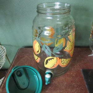 May include: A clear glass beverage dispenser featuring a colorful printed pattern of oranges and green leaves. The cylindrical container includes a white plastic spigot at the base and a separate dark green plastic lid with a circular opening.