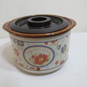 May include: A vintage potpourri crock with a black lid and floral design. The crock is off-white with a brown rim and features a central oval with the words "Potpourri Crock" and a red flower design. Blue and red flowers are scattered around the crock.