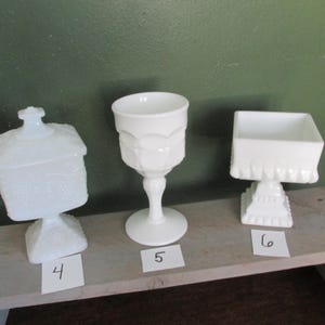 May include: Three white glass decorative pieces. The items include a lidded candy dish, a goblet, and a square dish. Each piece is on a white paper with a number. The background is a green wall.