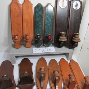 May include: A collection of wooden wall sconces in various styles and finishes. Some are stained in shades of brown, while others feature decorative elements like green marbleized panels and religious imagery. The sconces are designed to hold candles.