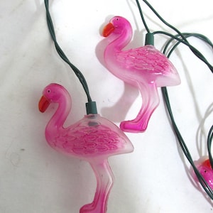 May include: Pink flamingo string lights with green wire. The lights are shaped like flamingos with pink bodies and red beaks.