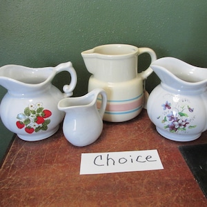 May include: A collection of ceramic pitchers in various sizes and designs. One pitcher features a strawberry motif, another has a floral pattern, and a third has pink and blue stripes. The word "Choice" is written on a white card.