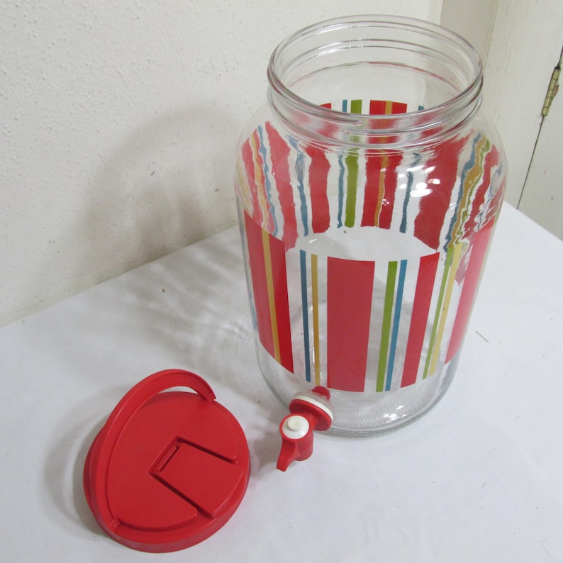 May include: A clear glass beverage dispenser featuring a vertical striped pattern in red, blue, and green. The dispenser includes a red plastic spigot at the base and a separate, matching red circular lid with a small flip-top opening.