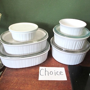 May include: A collection of white ceramic baking dishes with vertical ribbed sides, including oval and round shapes. Several dishes feature clear glass lids, and one small round dish is open. A white card with the handwritten word Choice is present.