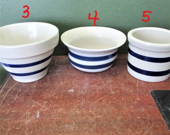 Peut inclure: Trois bols en céramique blanc cassé avec des rayures horizontales bleu foncé. Le bol marqué 3 a une base effilée, le bol marqué 4 est plus large avec un bord évasé, et le bol marqué 5 est cylindrique. Chaque bol porte des chiffres rouges.