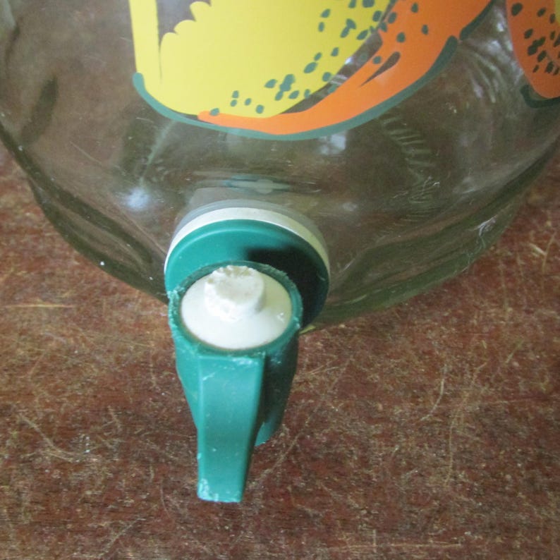 May include: A clear glass beverage dispenser featuring a green plastic spigot with a white push-button mechanism. The glass body displays a partial graphic print in yellow and orange tones, positioned above the spigot assembly on the container.