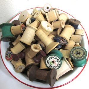 May include: A collection of vintage wooden spools in a white bowl with a red rim. The spools vary in color, including natural wood, dark brown, and green. Some spools have labels with text. A variety of sizes and shapes are visible.