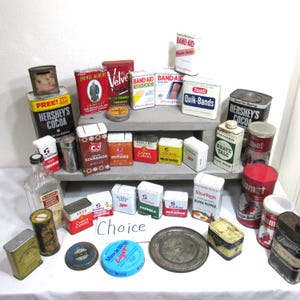 May include: A collection of vintage metal tins and containers, including Hershey's Cocoa, Band-Aid, and spice tins. The items are arranged on a white surface, with some displayed on a gray shelf. The word "Choice" is written on the surface.