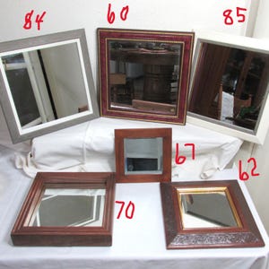 May include: A selection of square mirrors with diverse frame designs. The frames feature a grey wood effect, red and gold detailing, a white frame, dark wood, and a decorative brown frame with gold trim. The mirrors are arranged on a white surface.
